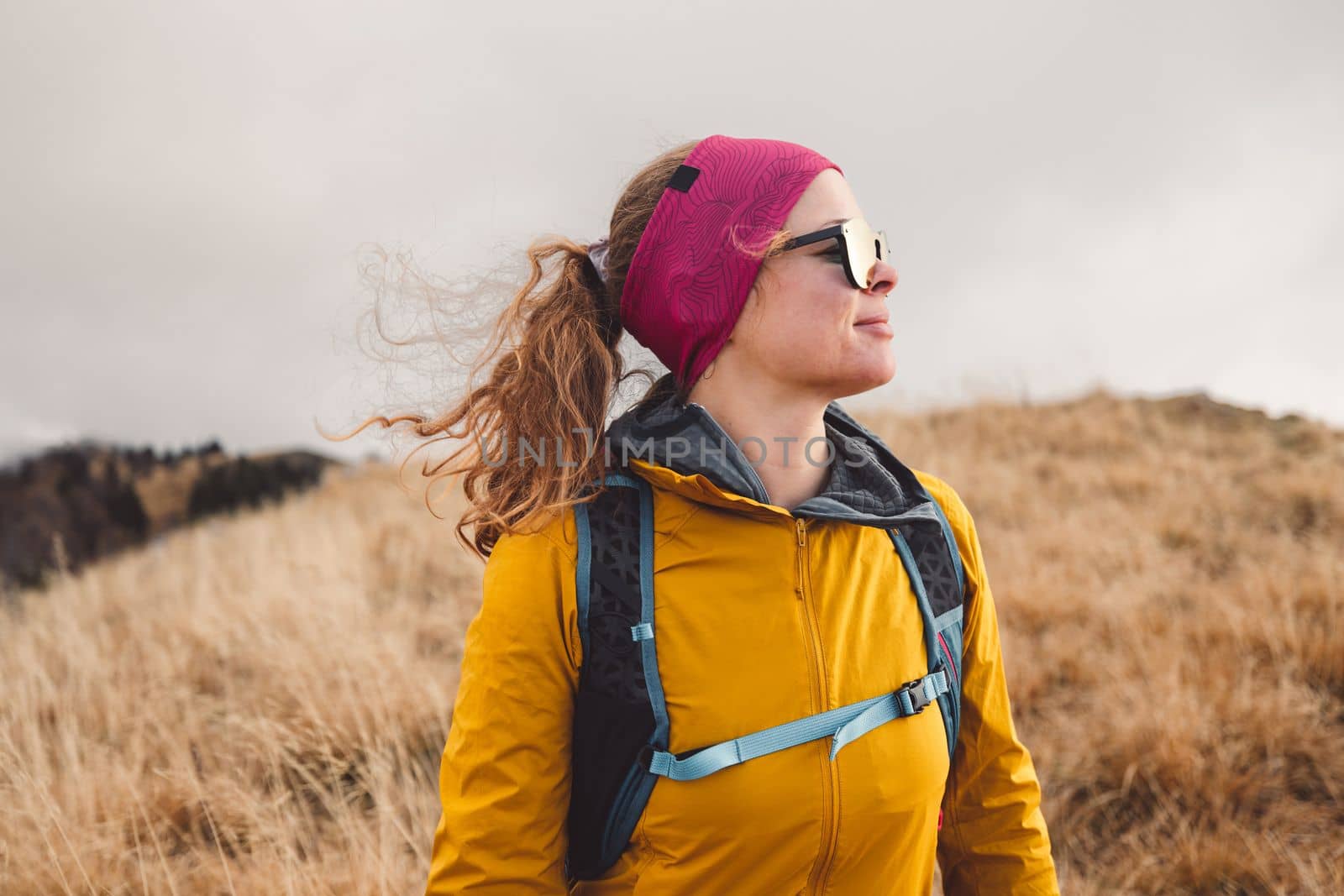 Caucasian woman hiker in yellow jacket and pink backpack hiking in the mountains on an autumn day, wearing gloves and headband. 