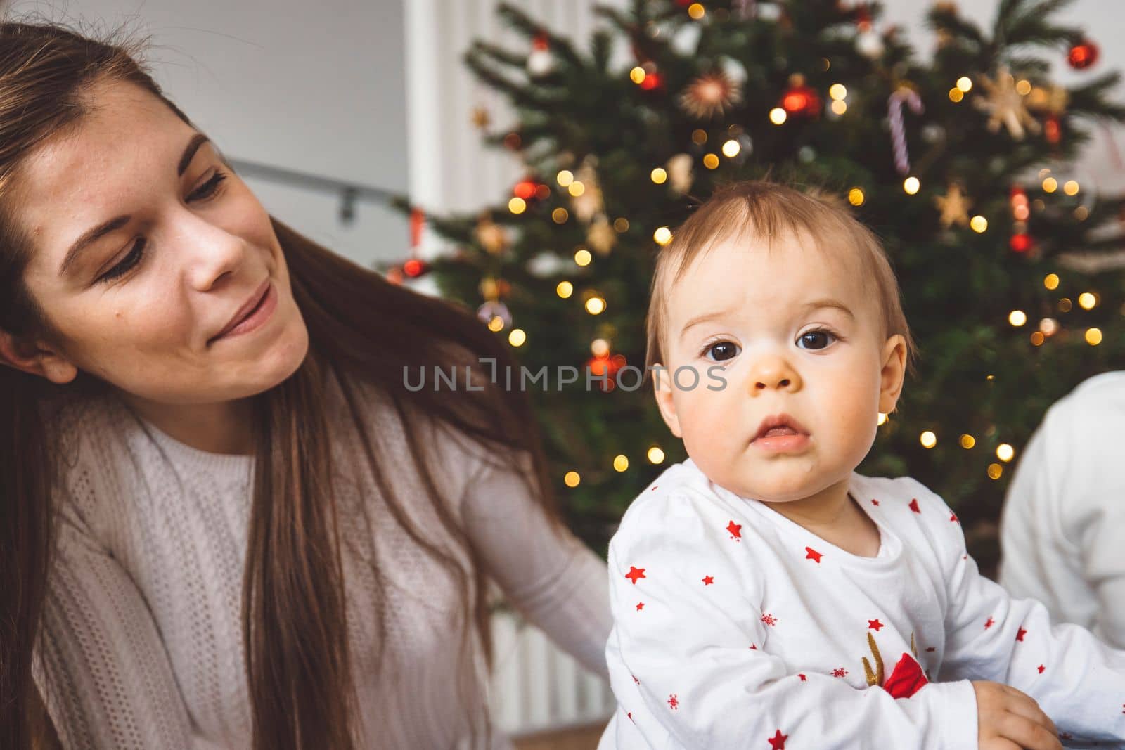 Baby girl looking in the camera while her mom watcher her, Christmas tree in the background by VisualProductions