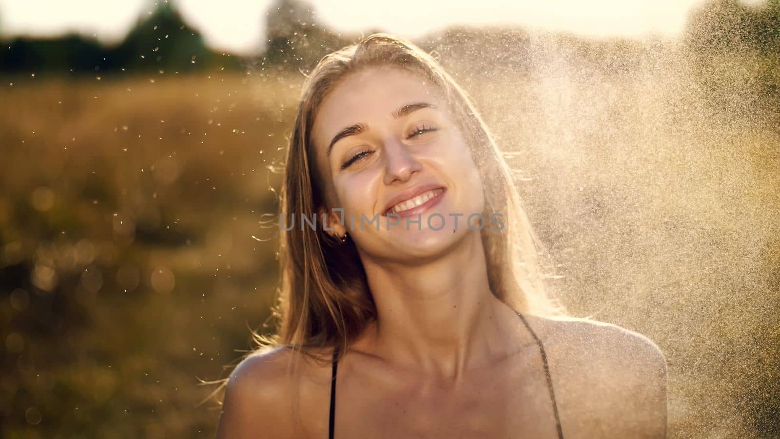 Portrait of a beautiful young blonde girl, under light summer rain, in the sun rays, on a green meadow. The girl smiles, strokes herself over the delicate, beautiful white skin. High quality photo