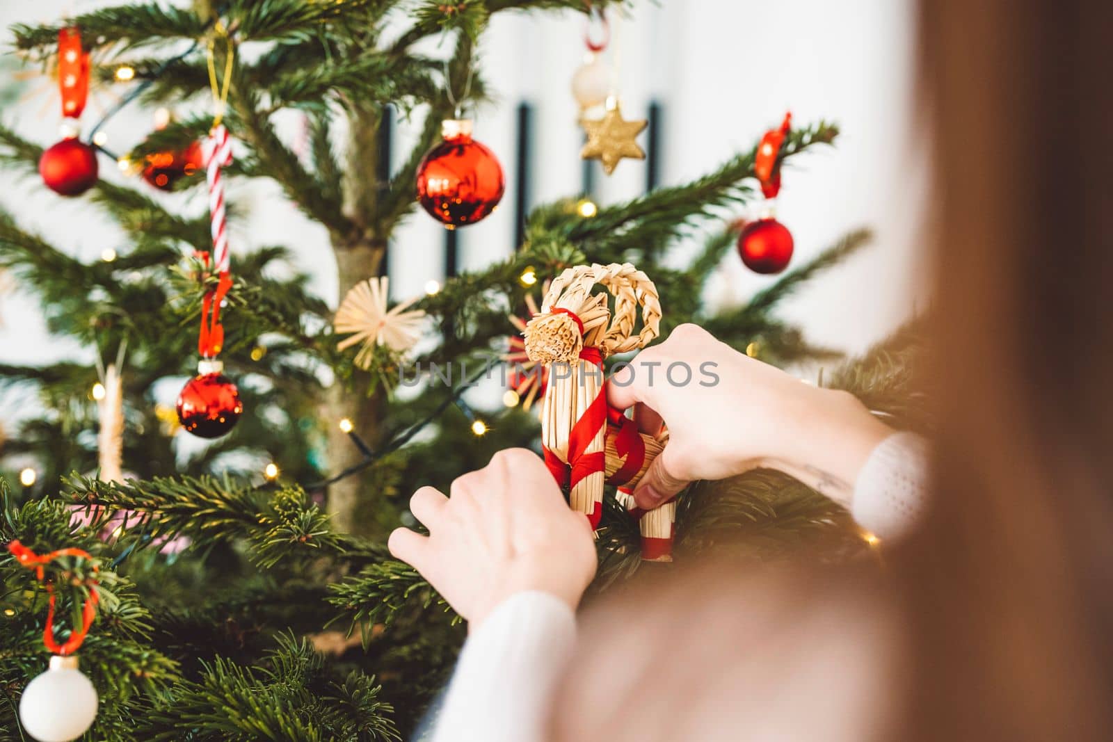 Unrecognizable caucasian woman decorating the Christmas tree, putting red ornament on the tree, holding a red Christmas ornament. 