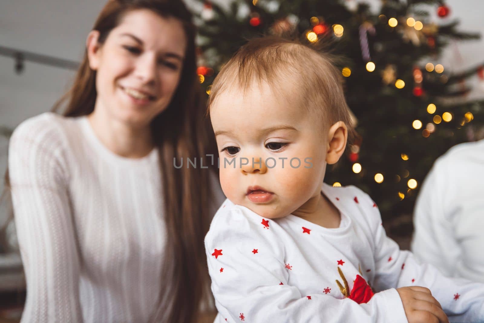 Portrait of young mom and her daughter, baby girl having fun on Christmas, decorating the Christmas tree in festive outfits. Smiling baby girl playing with her mom. 