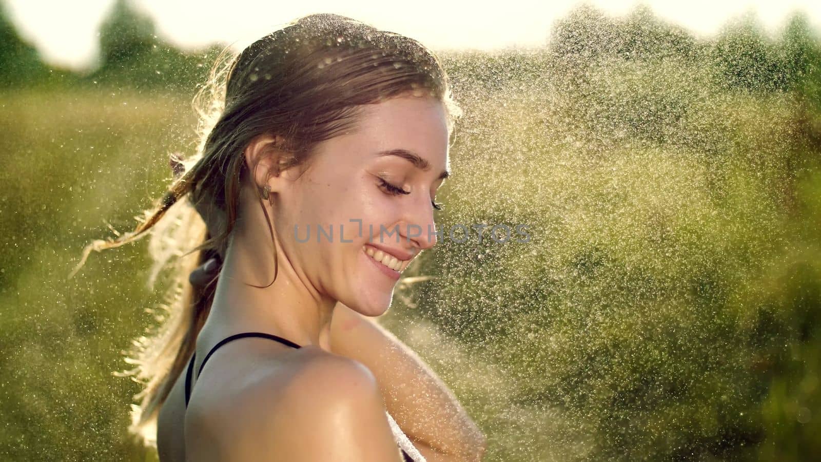 Portrait of a sexy, beautiful young blonde girl, under a light summer rain, in the sun rays, on a green meadow. The girl smiles, Her skin shines, radiates health. Image of happiness. High quality photo