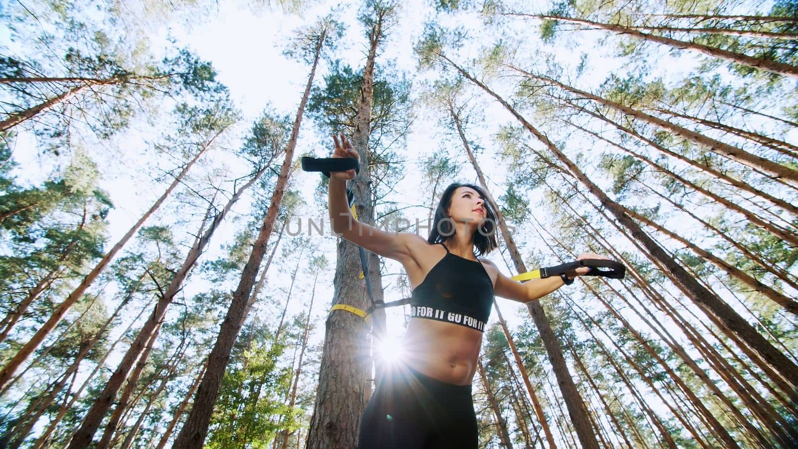 athletic woman, coach, instructor, performs, doing exercises. In pine forest, in summer, in sun rays. View from below, bottom view by djtreneryay