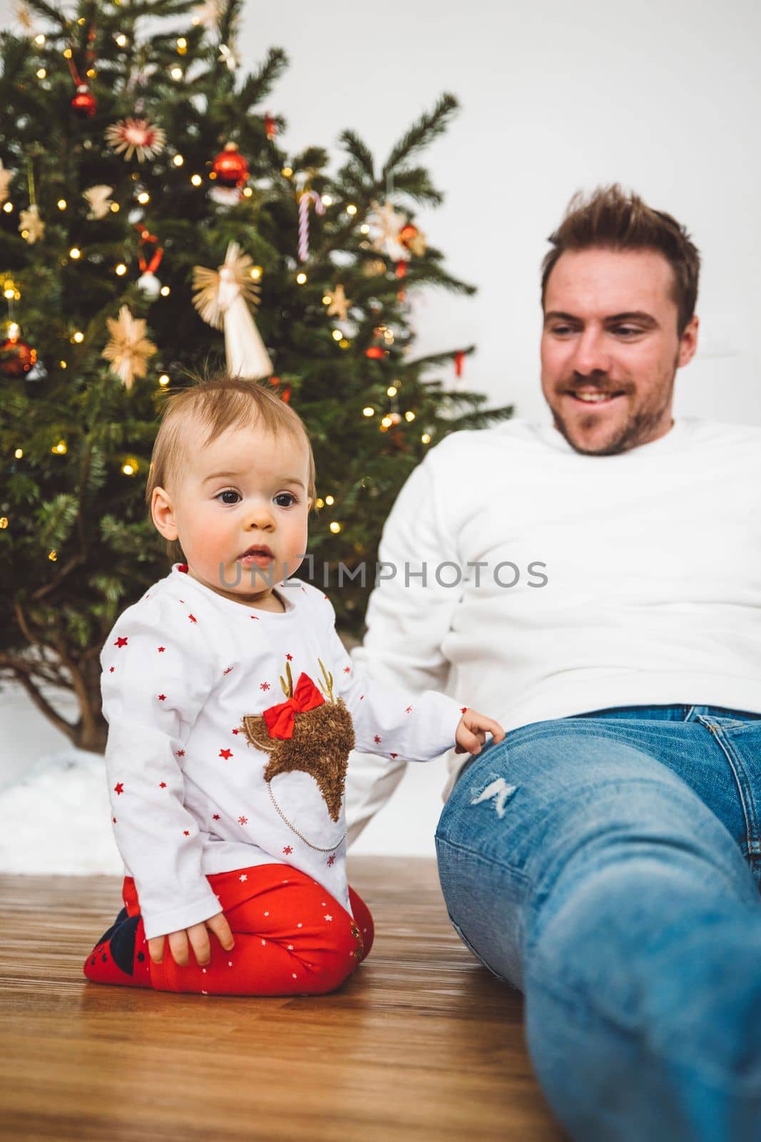 Father and daughter, baby girl having fun on Christmas, decorating the Christmass tree in festive outfits. Smiling baby girl playing with her dad. 