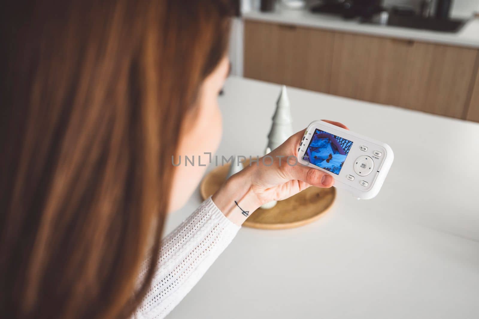 Young caucasian woman dressed in white sitting by the white kitchen island looking at the baby monitor, watching her baby sleep. 