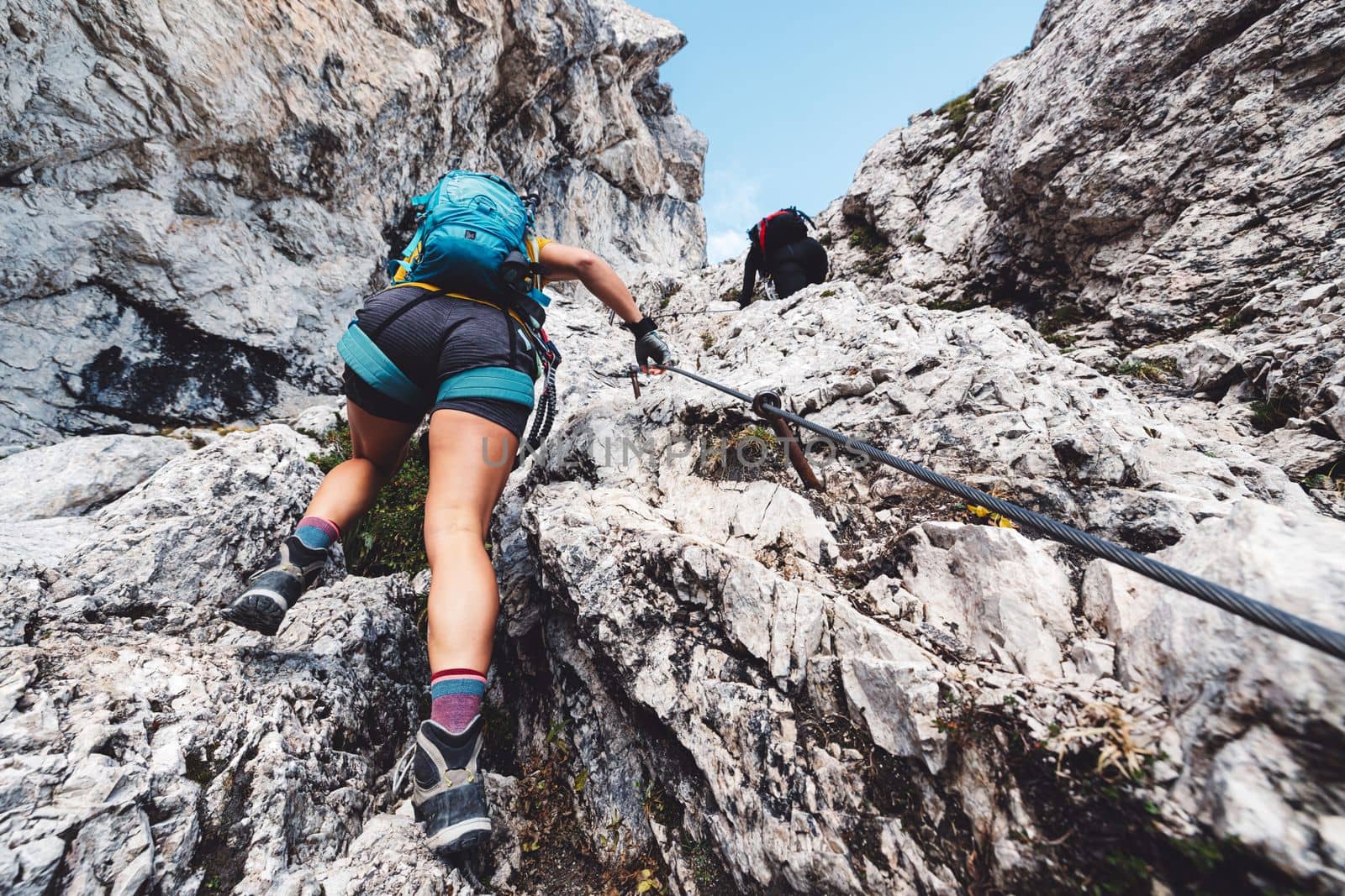 Caucasian woman mountaineer climbing up the mountain on via ferrata trail, high up in the mountain peaks. Woman wearing a protective head gear and uses a safety harness while she climbs.