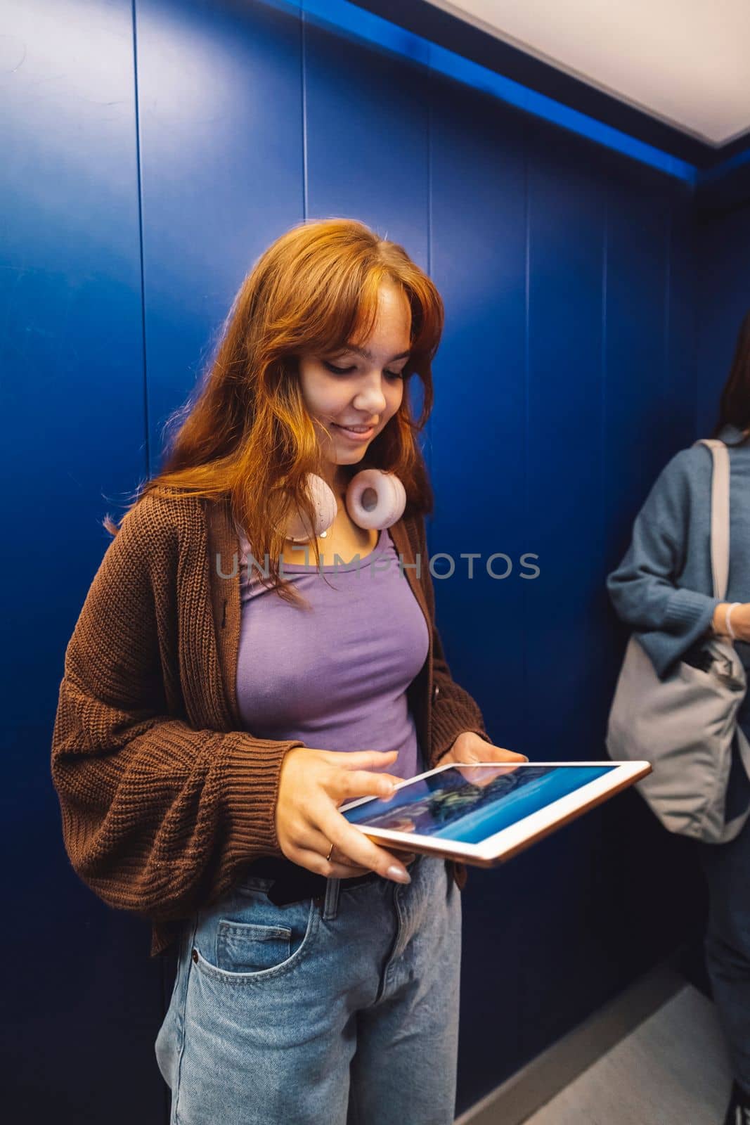 Vertical photo of young caucasian woman with handphones around her neck, holding a dgital tablet while standing in an elevator.