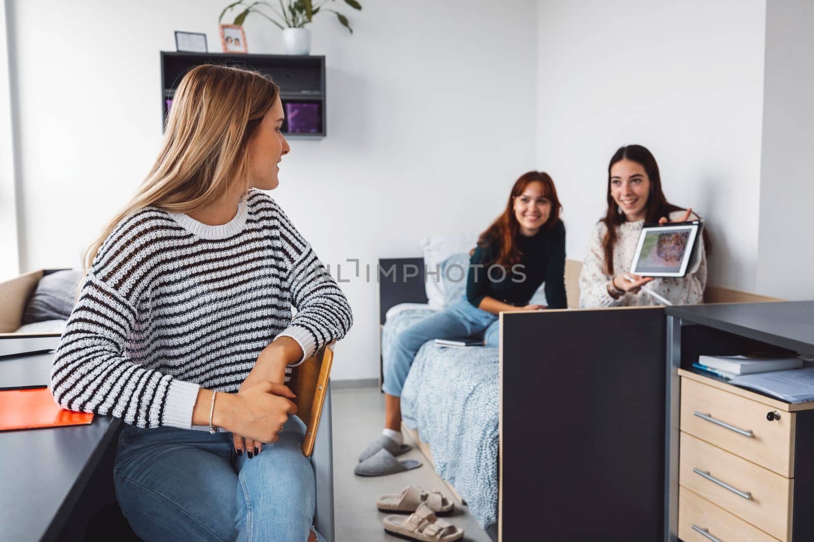 College students in their dorm room, one sitting on the chair, looking back at their friends, showing her something on the tablet by VisualProductions