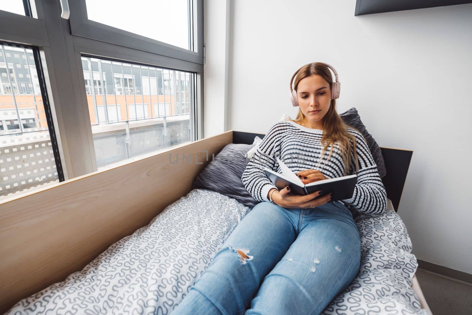 Blonde young caucasian woman lying on her dorm room bed next to a window putting on headphones, listening to music on a cold cloudy autumn day.