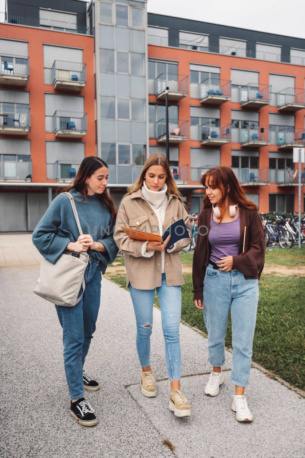 Group of three cheerful female collage students outside their dorm on a cold autumn day, walking on campus to get to their class. 