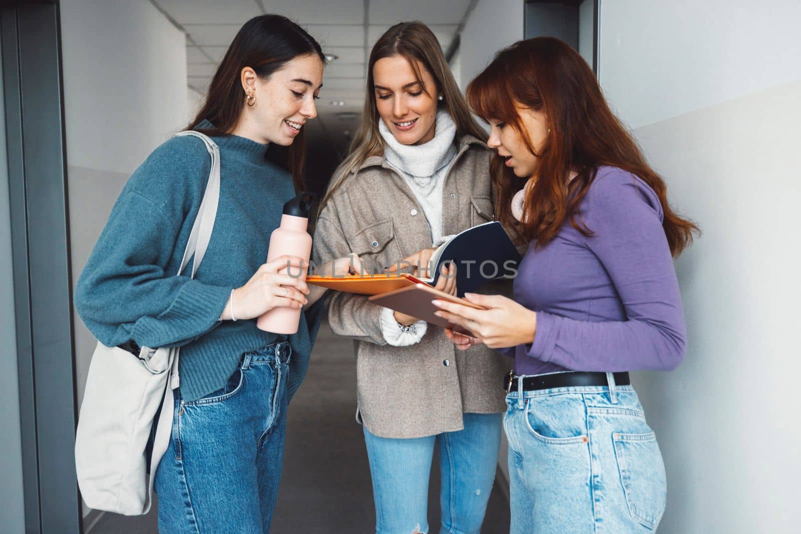 Group of three friends, young caucasian women students walking inside the school during a break, headed to another classroom. Students carrying their books with them. 