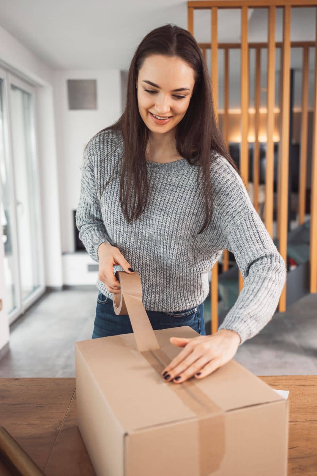 Vertical photo of young woman sending Christmas presents to family, taping a brown cardboard box by VisualProductions
