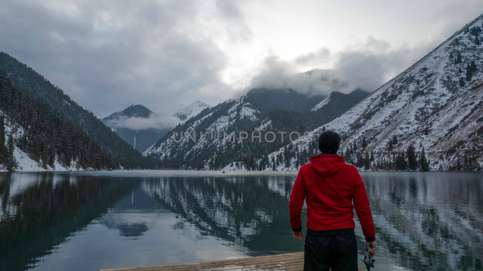 The guy standing on the pier admires the mountain lake. There is a view of the mirrored black color of the water, which reflects snowy mountains, green forest, clouds and a yellow sunset. Kolsai Lake