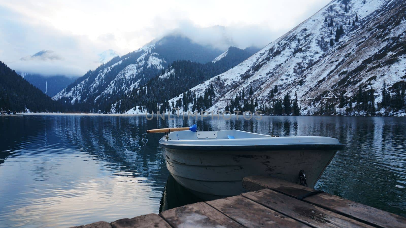 A mountain lake in the forest with mirrored water. Wooden pier with white boat. The water reflects the landscape of a cloudy sky, snowy mountains and peaks, coniferous firs. Kolsai Lake, Kazakhstan