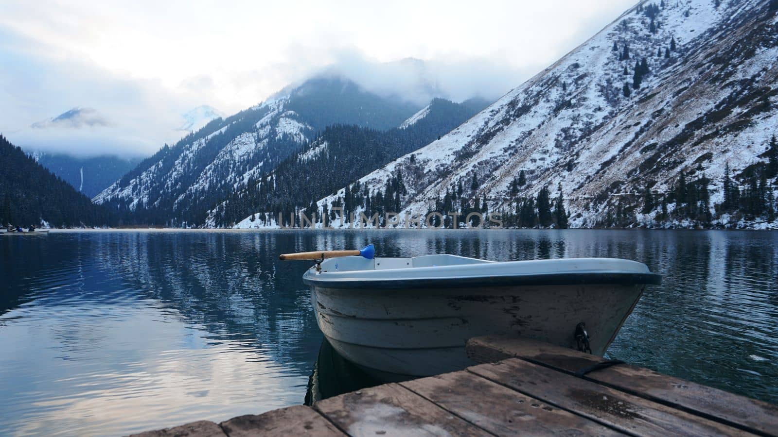 A mountain lake in the forest with mirrored water. Wooden pier with white boat. The water reflects the landscape of a cloudy sky, snowy mountains and peaks, coniferous firs. Kolsai Lake, Kazakhstan