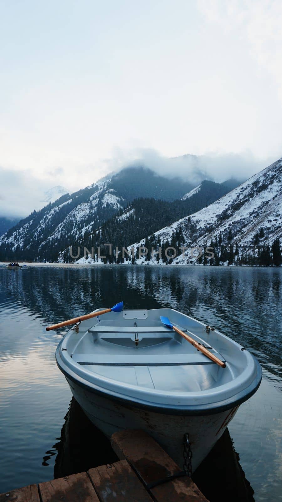 A mountain lake in the forest with mirrored water. Wooden pier with white boats. The water reflects the landscape of a cloudy sky, snowy mountains and peaks, coniferous firs. Kolsai Lake, Kazakhstan