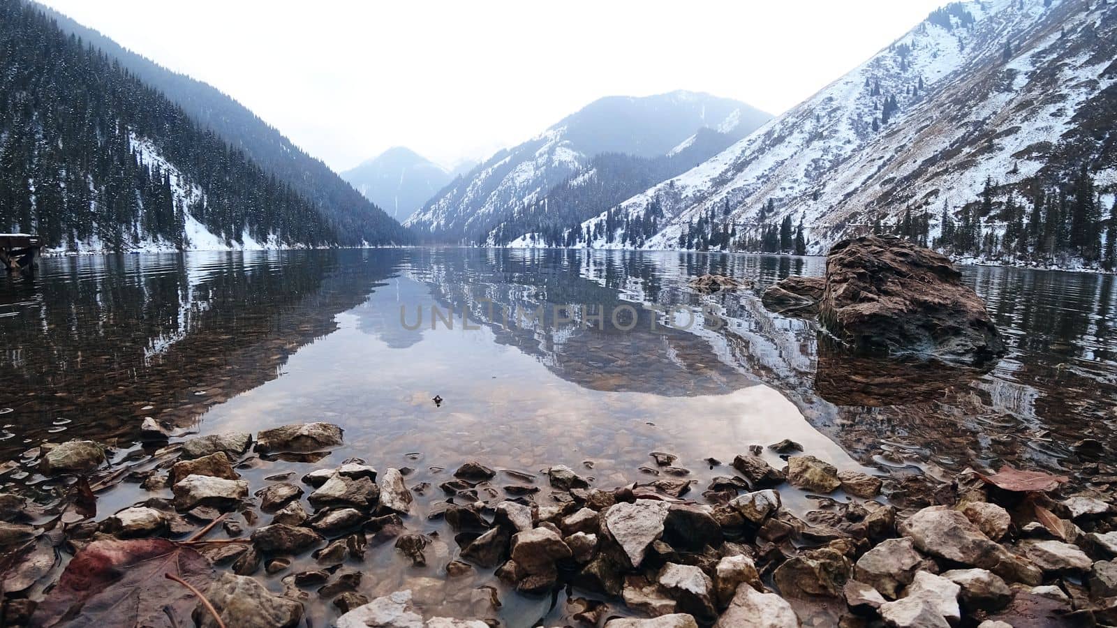 Kolsai mountain lake in the winter forest. View of clouds, coniferous trees, mirror-smooth water, hills and mountains in the snow. The landscape is reflected in the water. There are stones and leaves