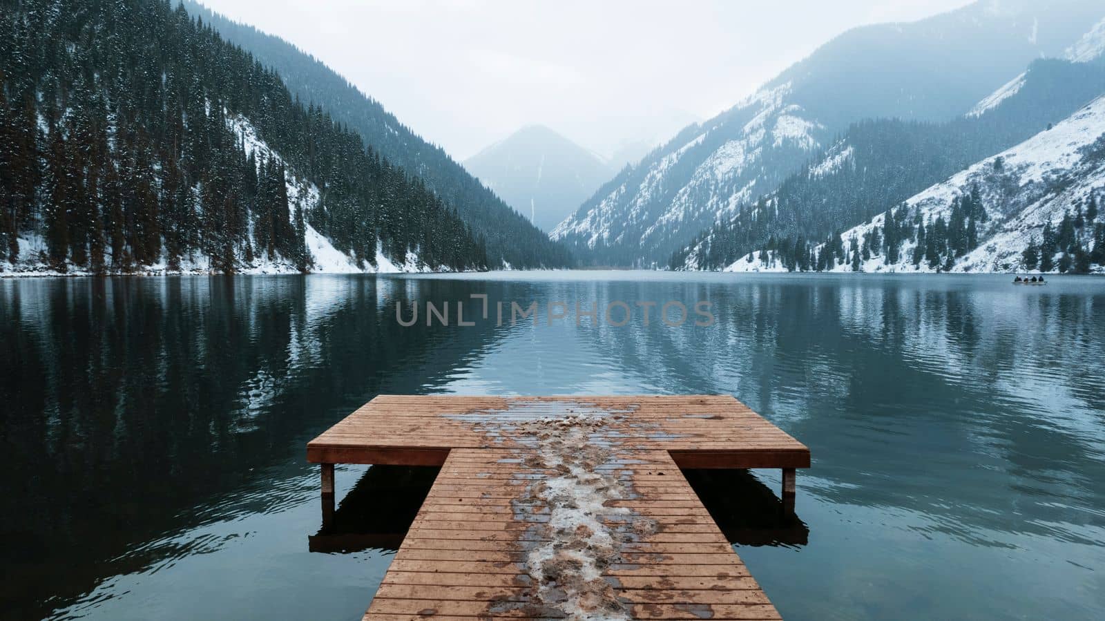 Kolsai mountain lake in the winter forest. Drone view of a wooden pier, white clouds, coniferous trees, mirror-smooth water, hills and mountains in the snow. Boats float in places. Kazakhstan, Almaty