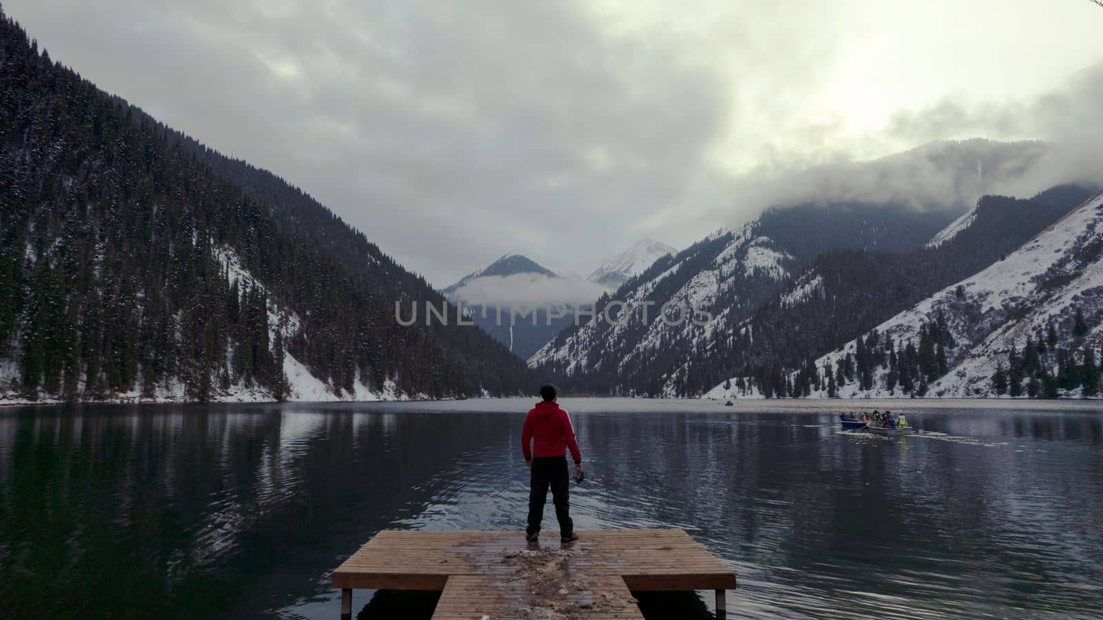 The guy standing on the pier admires the mountain lake. There is a view of the mirrored black color of the water, which reflects snowy mountains, green forest, clouds and a yellow sunset. Kolsai Lake