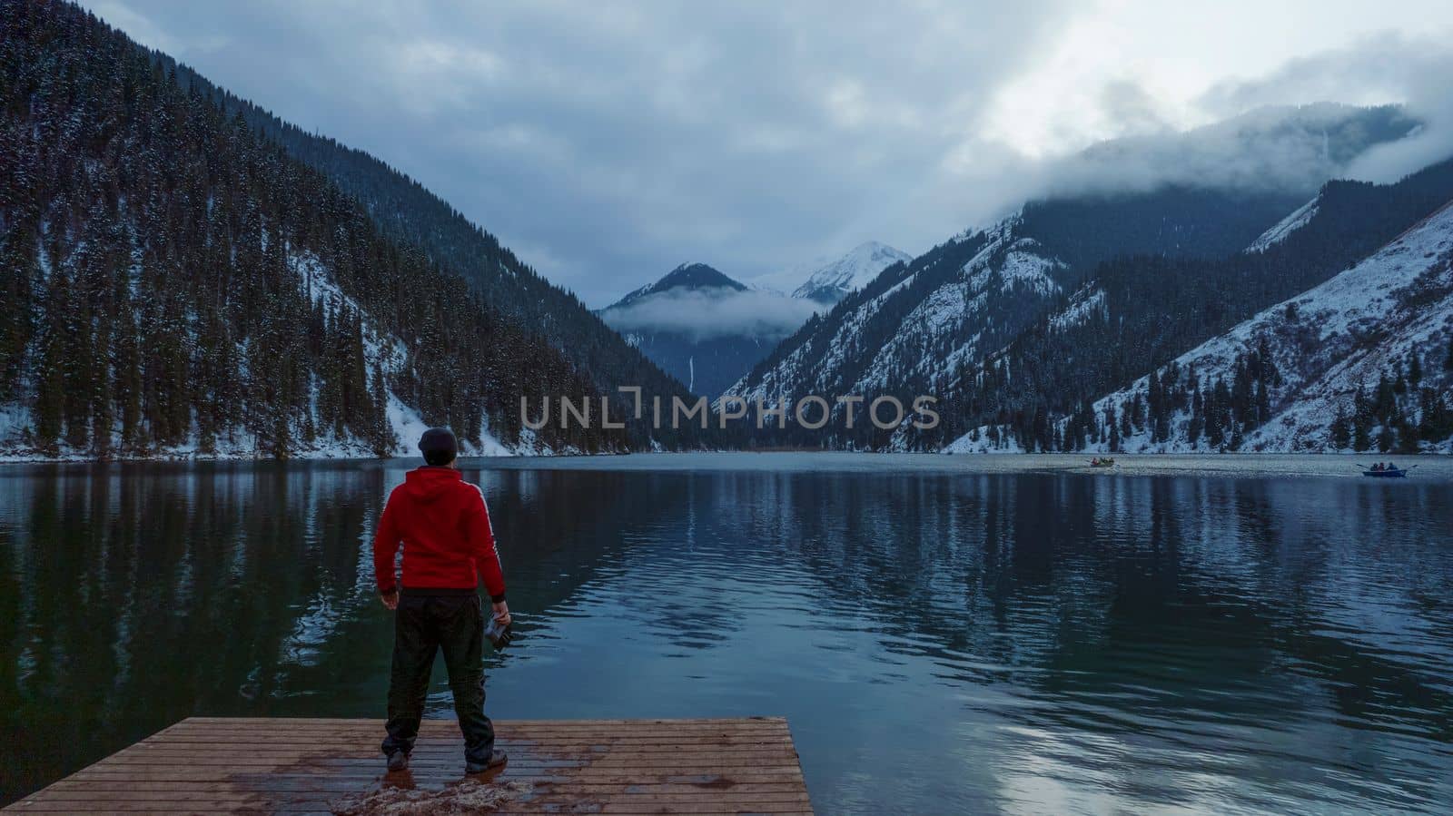 The guy standing on the pier admires the mountain lake. There is a view of the mirrored black color of the water, which reflects snowy mountains, green forest, clouds and a yellow sunset. Kolsai Lake