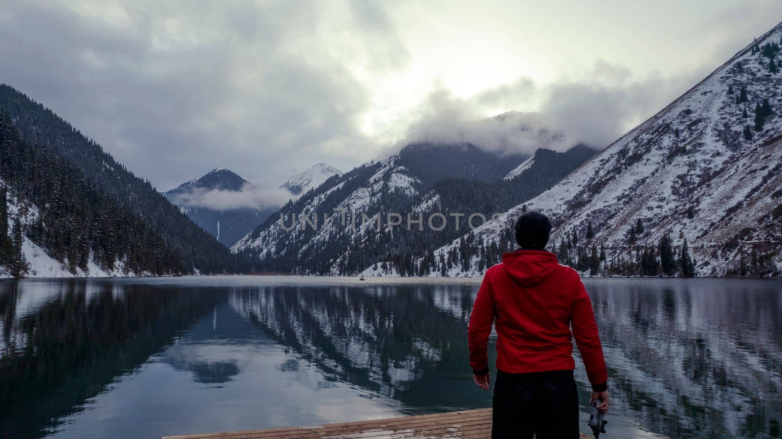 The guy standing on the pier admires the mountain lake. There is a view of the mirrored black color of the water, which reflects snowy mountains, green forest, clouds and a yellow sunset. Kolsai Lake