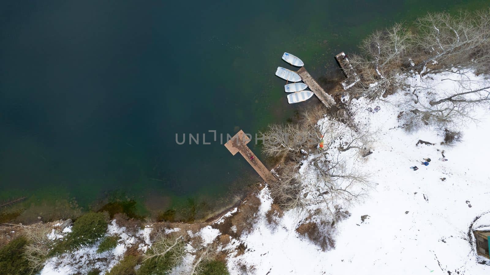 Kolsai mountain lake in the winter forest. Drone view of the pier with boats, trees, clear smooth water, snow lying on the ground. Rocks are visible in the water. Kazakhstan, Almaty
