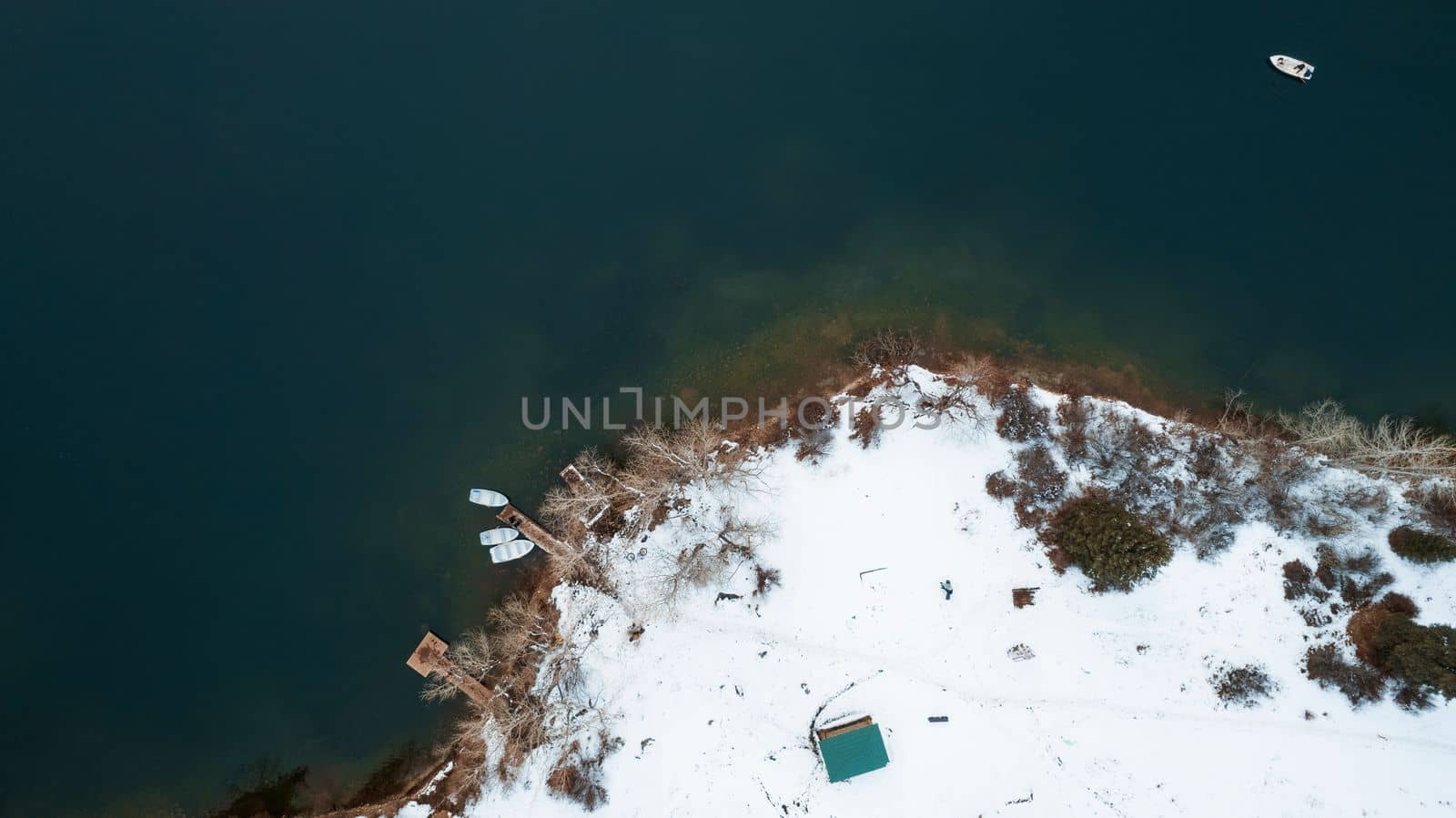Kolsai mountain lake in the winter forest. Drone view of the pier with boats, trees, clear smooth water, snow lying on the ground. People swim in white boats. Kazakhstan, Almaty