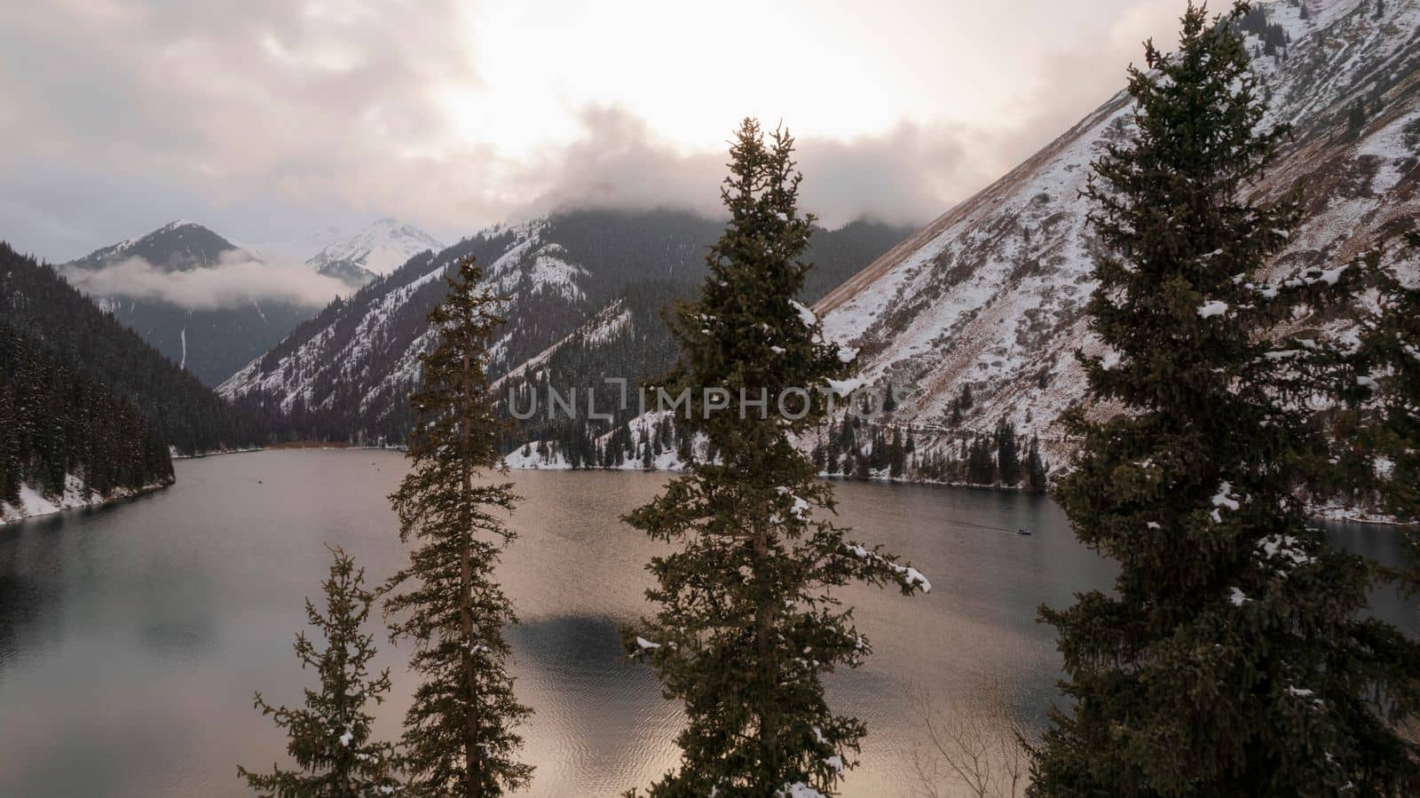 Kolsai mountain lake in the winter forest. Drone view of coniferous trees, mirror-smooth water like a mirror, hills, mountains in snow and clouds. Yellow sunset. Boats float in places. Kazakhstan