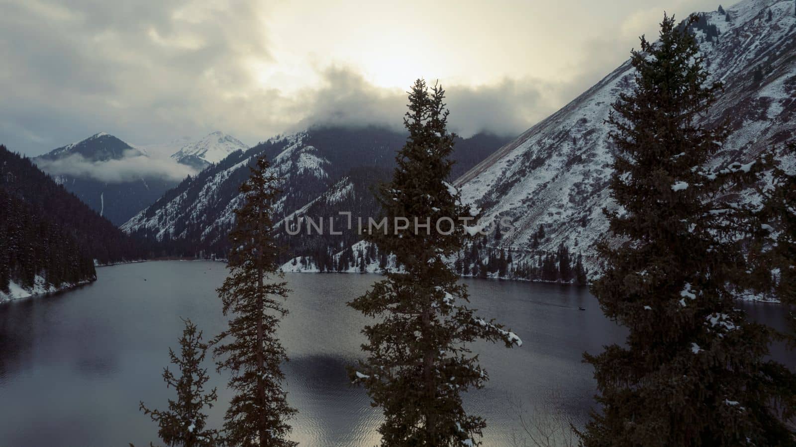 Kolsai mountain lake in the winter forest. Drone view of coniferous trees, mirror-smooth water like a mirror, hills, mountains in snow and clouds. Yellow sunset. Boats float in places. Kazakhstan