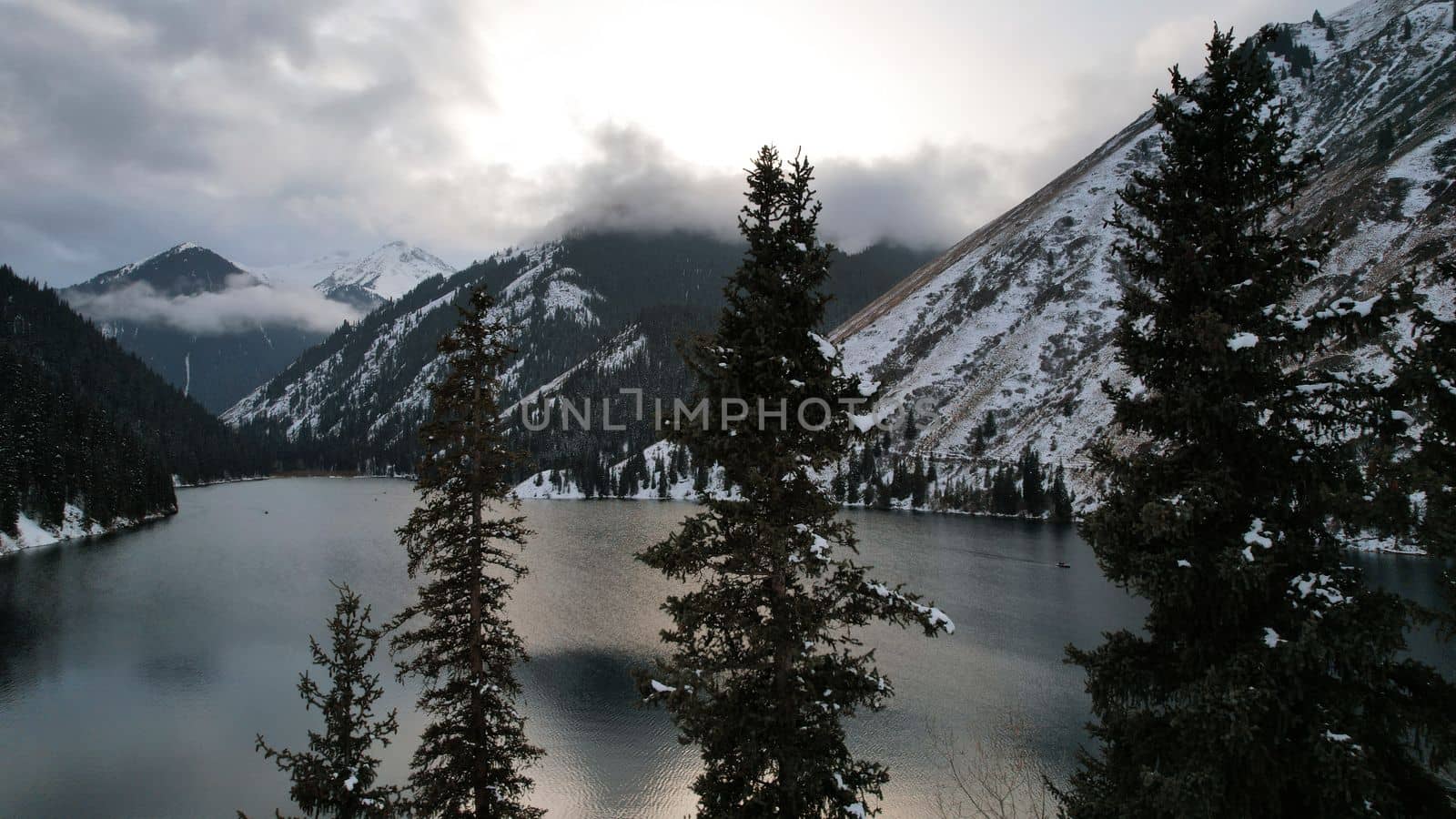 Kolsai mountain lake in the winter forest. Drone view of coniferous trees, mirror-smooth water like a mirror, hills, mountains in snow and clouds. Yellow sunset. Boats float in places. Kazakhstan