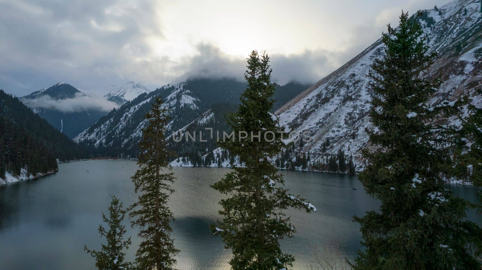 Kolsai mountain lake in the winter forest. Drone view of coniferous trees, mirror-smooth water like a mirror, hills, mountains in snow and clouds. Yellow sunset. Boats float in places. Kazakhstan