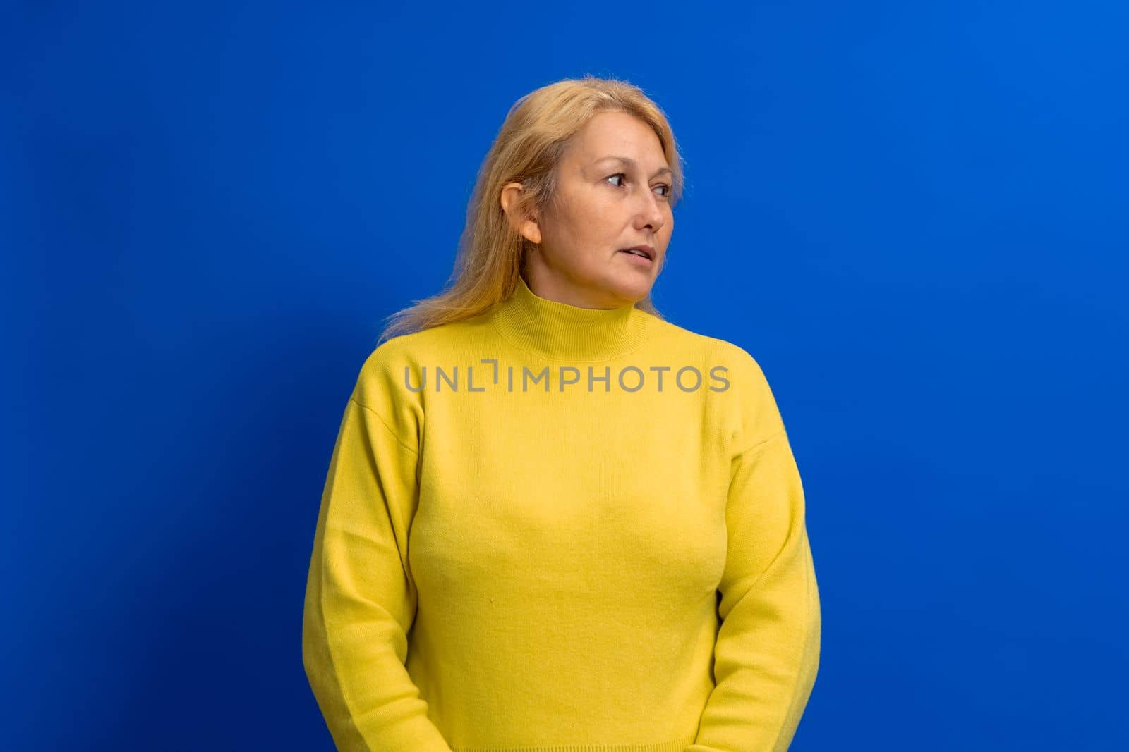Beautiful woman in her 40s wearing yellow sweater standing on blue background looking to side with serious face