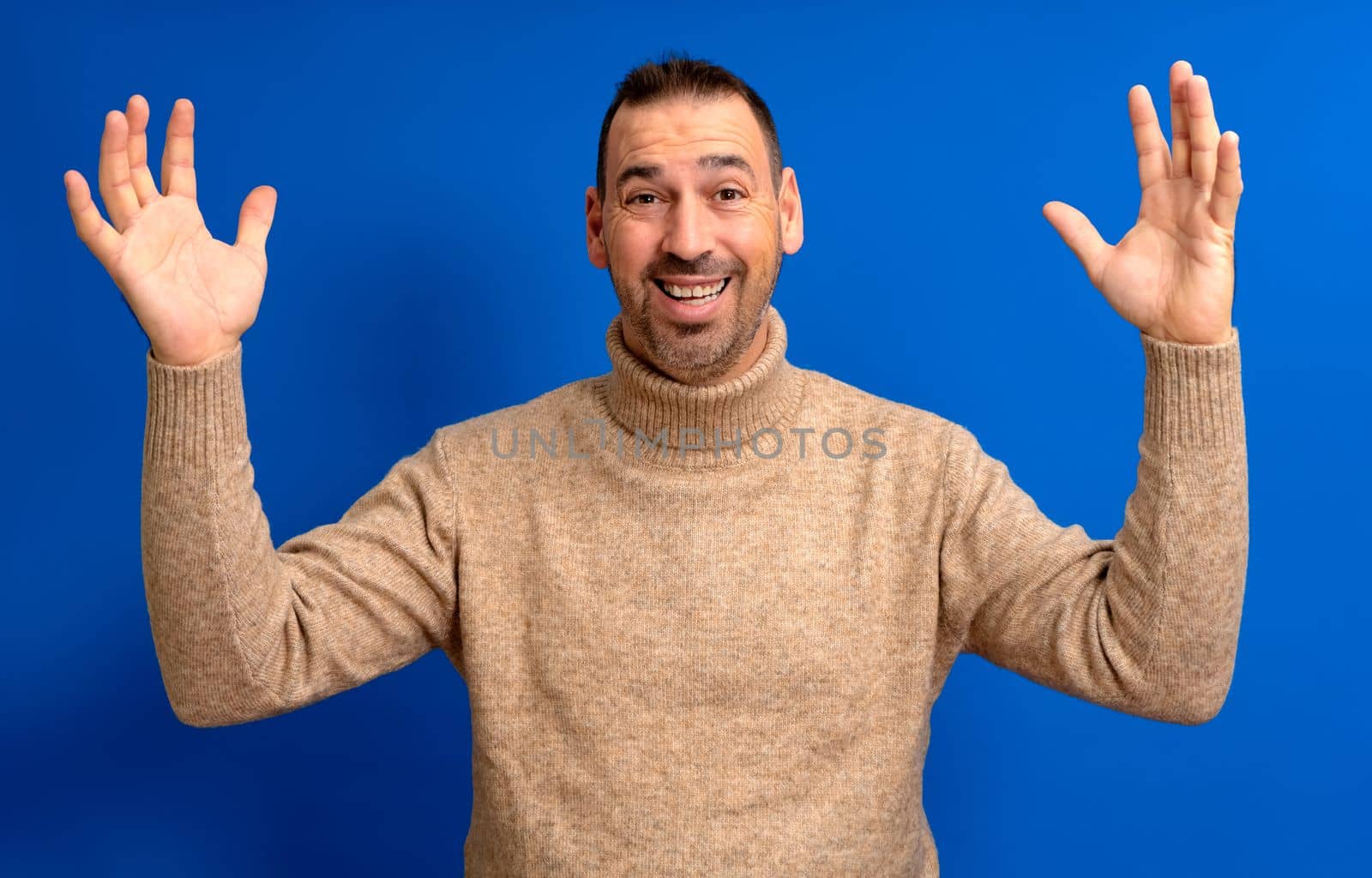 Smiling happy man in his 40s in a turtleneck holds his arms up in an expression of joy on a blue background studio portrait. People lifestyle concept