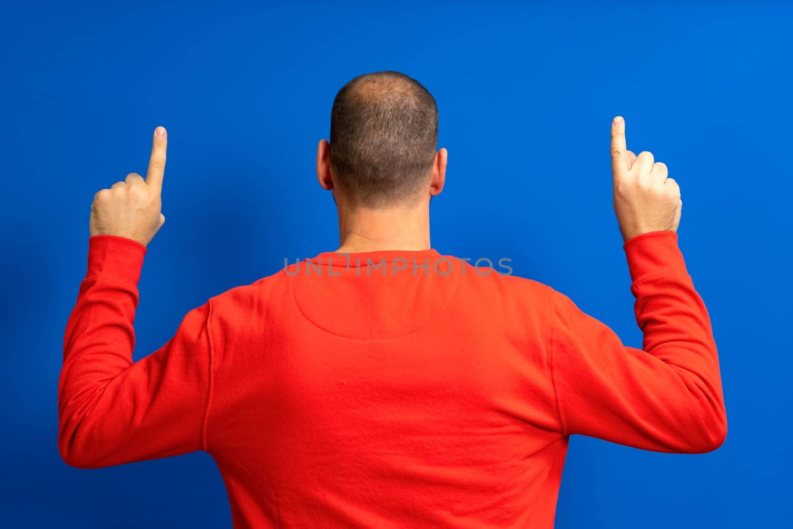 Portrait of a man with incipient alopecia from behind, raising both hands and pointing his index fingers up, isolated on a blue background