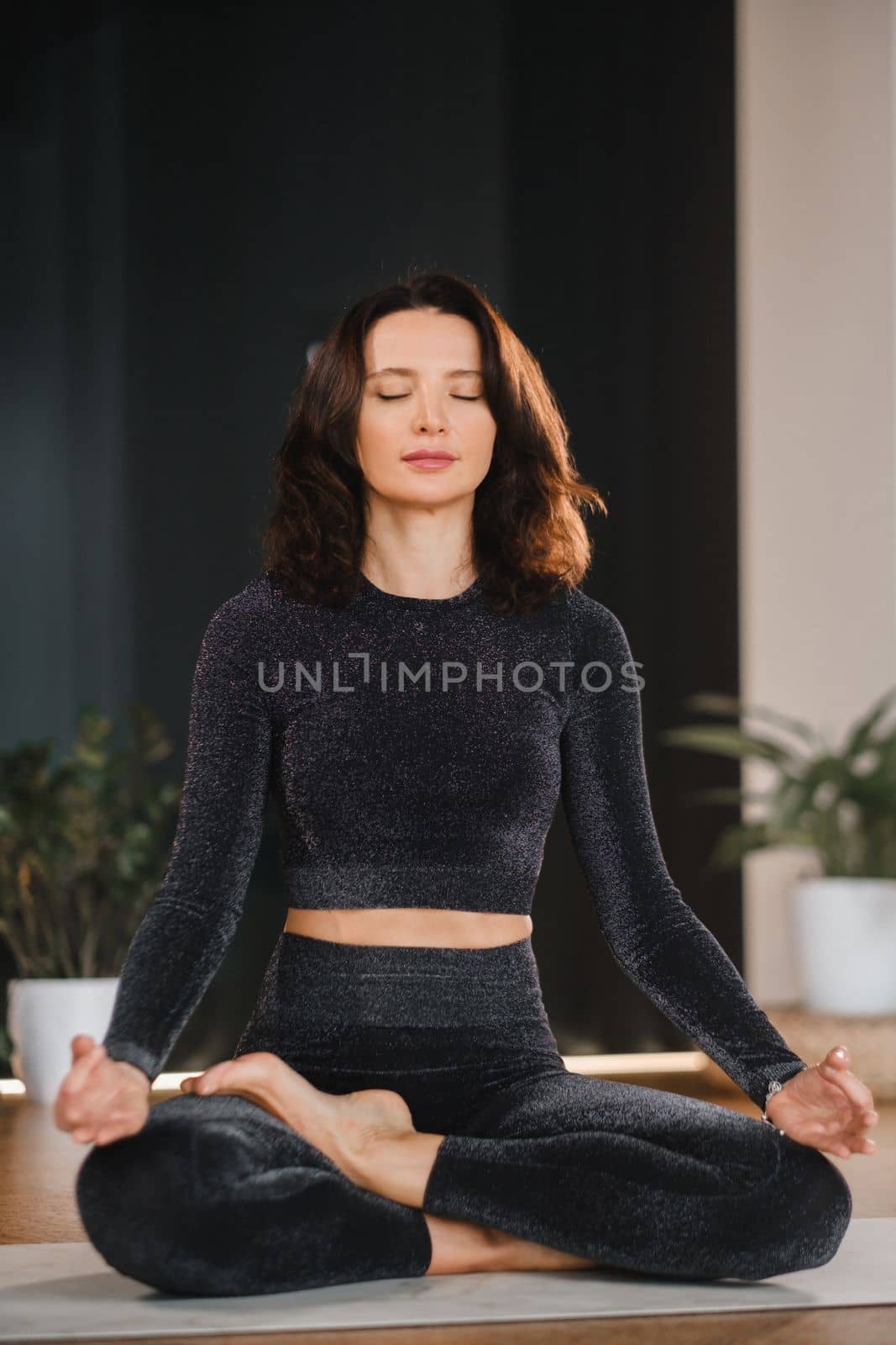A woman in a black tracksuit sits on a yoga mat in a lotus position in the gym by Lobachad