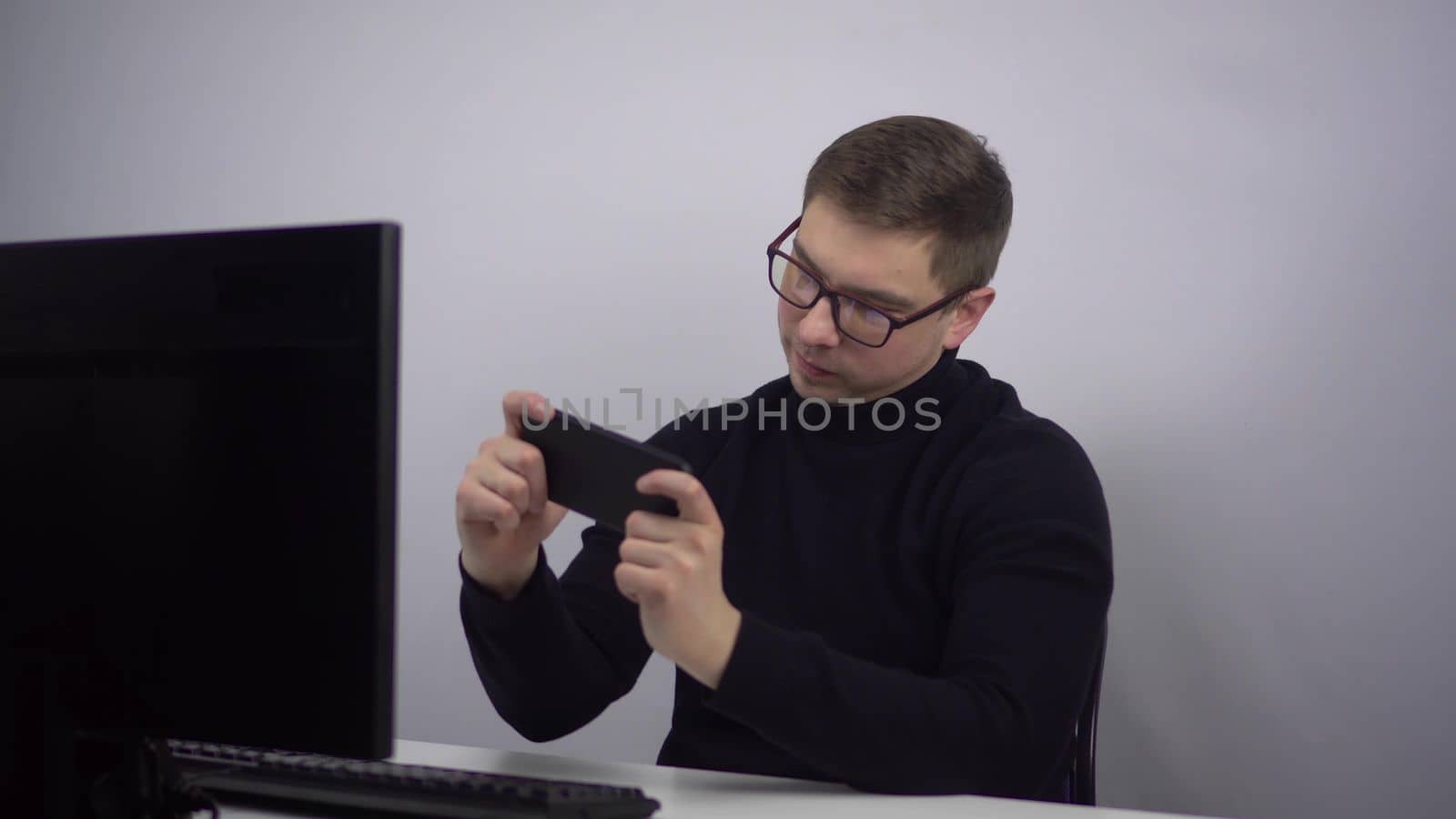 The young man is enthusiastically playing his smartphone and emotionally rejoices at the victory. IT specialist in glasses with a phone in his hands at the workplace in the office. 4k