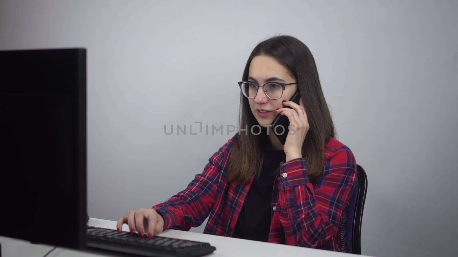 IT specialist sits at the workplace in the office and speaks on the phone. A young woman in glasses and a red shirt is working. 4k