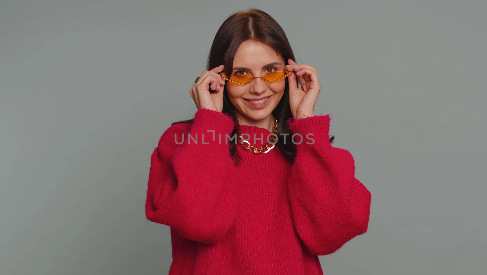 Portrait of happy pretty young woman in sweater wearing sunglasses, looking at camera with toothy charming smile, flirting, expressing optimism. Millennial girl isolated on gray studio background