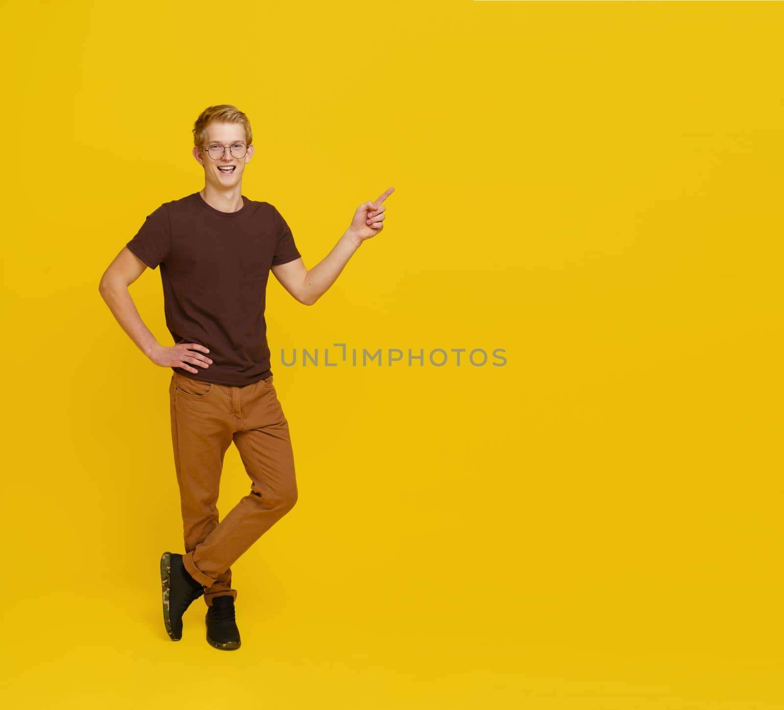 Young Successful Guy Appoints To Empty Place for Copy Space. Man 20s in brown t-shirt in foxy jeans Smiling Isolated on Yellow Background. High quality photo