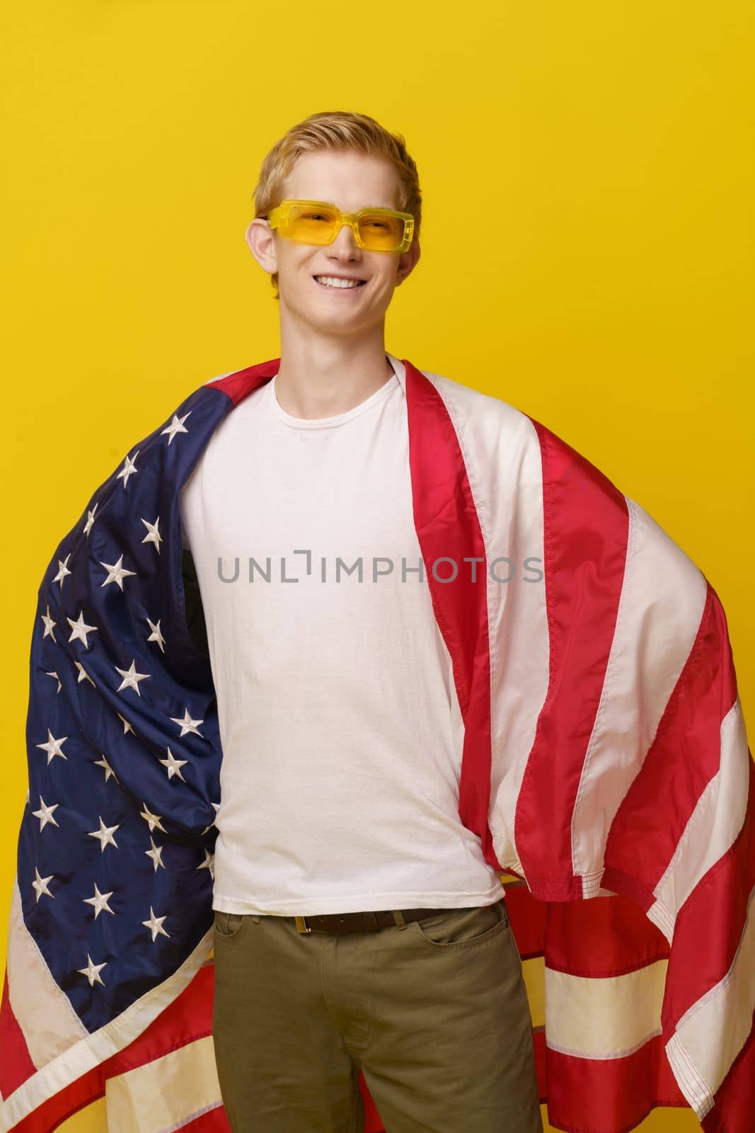 Super American Hero Concept. Youth Smiles in a White T-shirt and the American Flag is isolated on a yellow background. by LipikStockMedia
