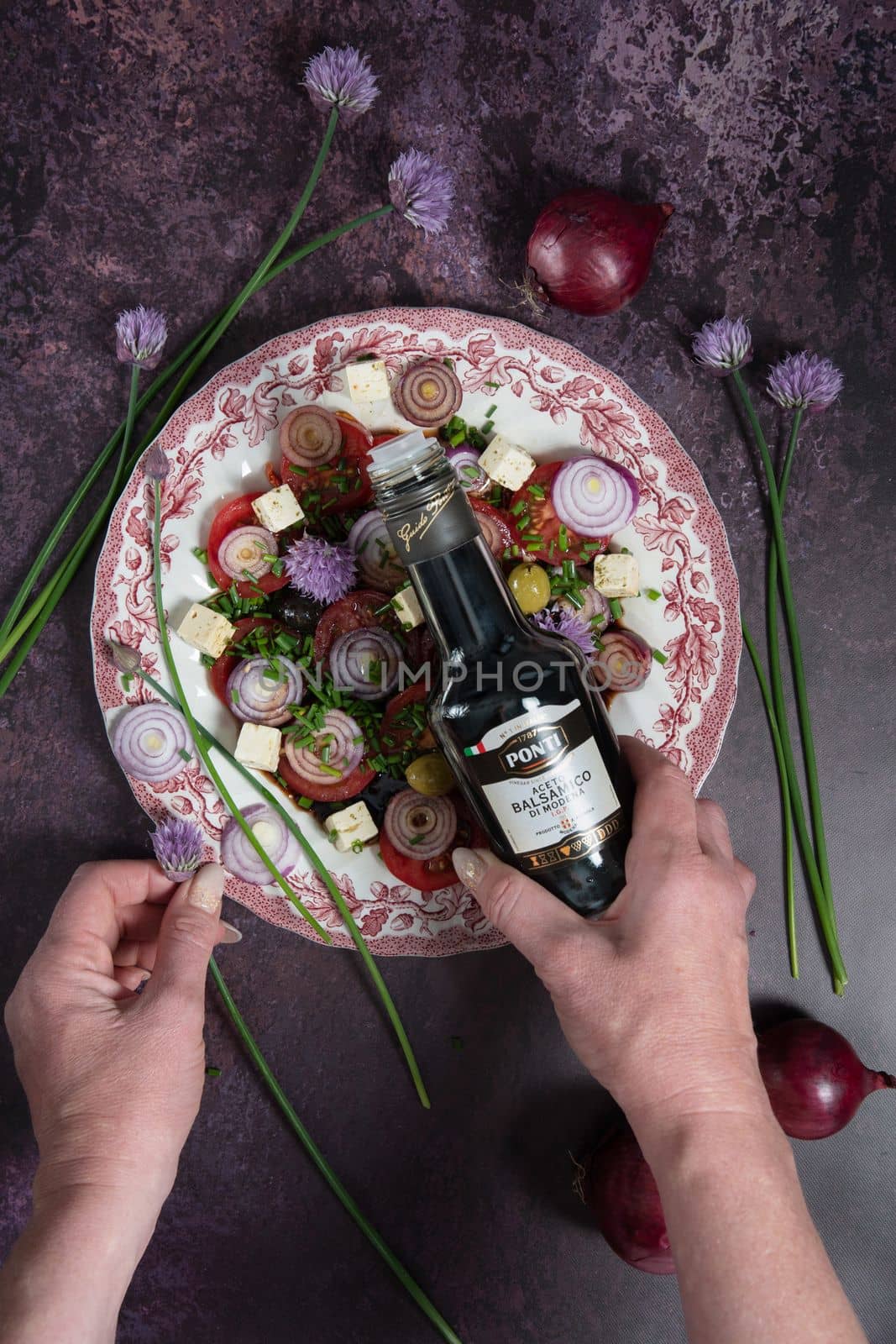 As,Belgium, May 10, 2022: tomato salad with purple onion rings dressed with Ponti balsamic dressing and olive oil with feta cheese and olives on a dark purple background, step by step recipe,flat lay