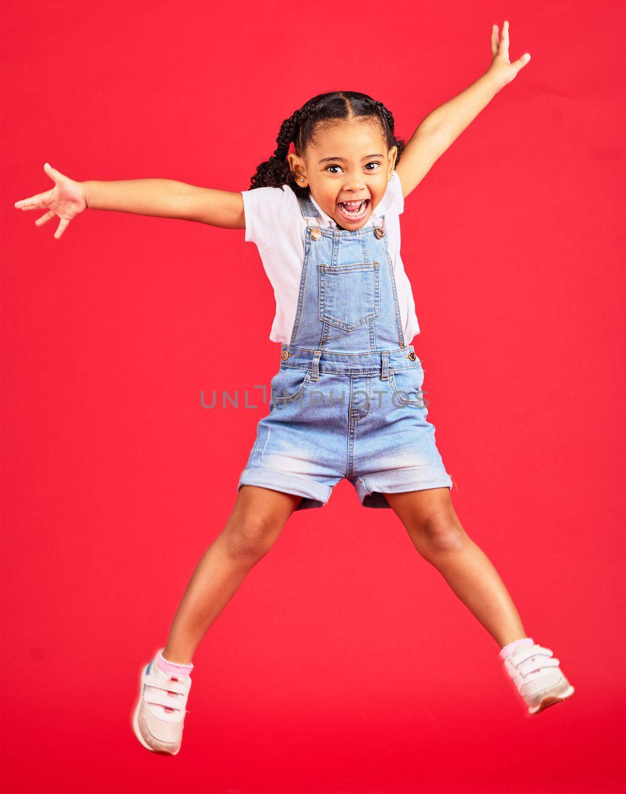 Excited, playful and portrait of a girl jumping while isolated on a red background in a studio. Youth, smile and child with freedom, energy and happiness while playing with a jump on a backdrop.