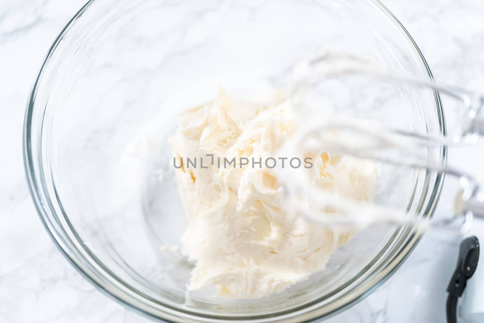 Mixing store-bought vanilla frosting in a mixing glass bowl with a hand mixer.