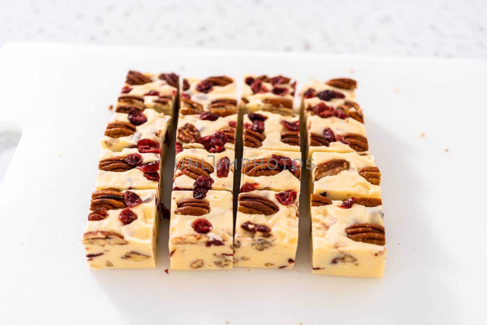 Cutting white chocolate cranberry pecan fudge into small pieces on a white cutting board.