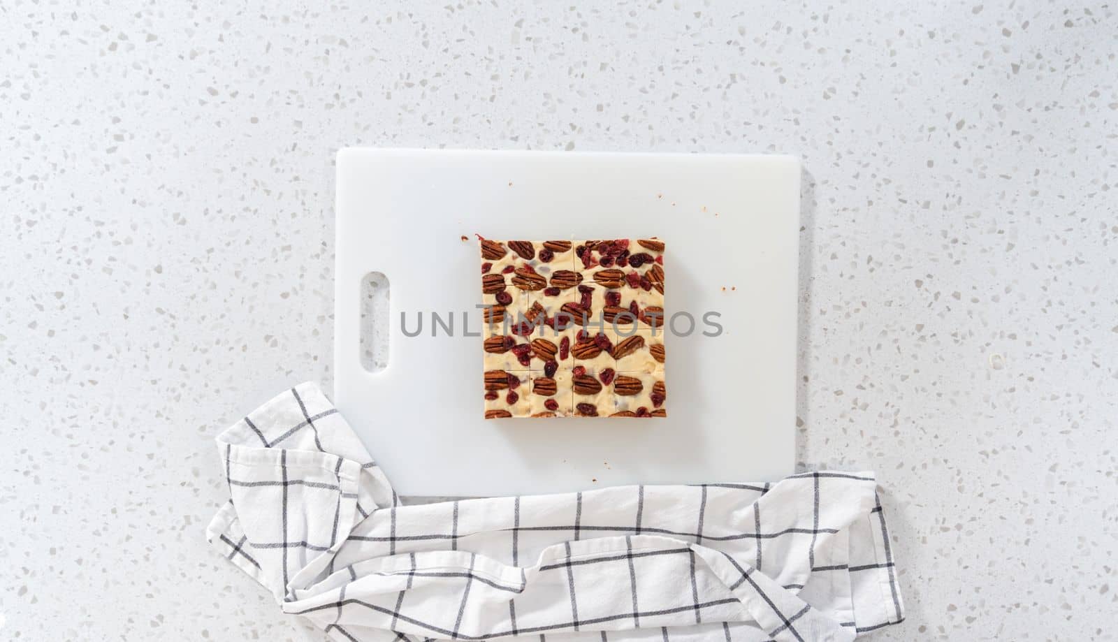 Flat lay. Cutting white chocolate cranberry pecan fudge into small pieces on a white cutting board.