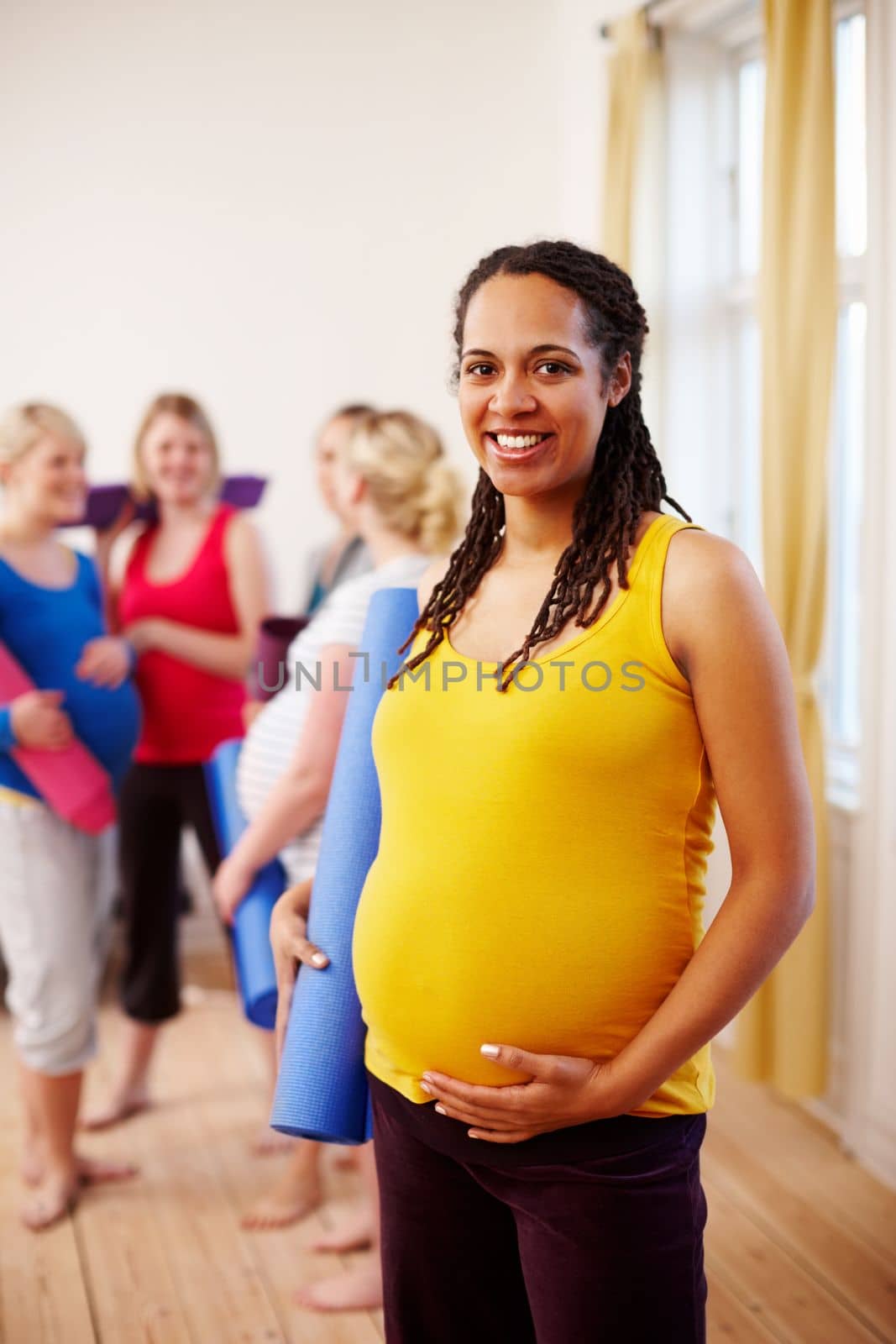 Enjoying a healthy pregnancy lifestyle. A happy young pregnant African-American woman holding her exercise mat while standing in a gym with her friends in the background