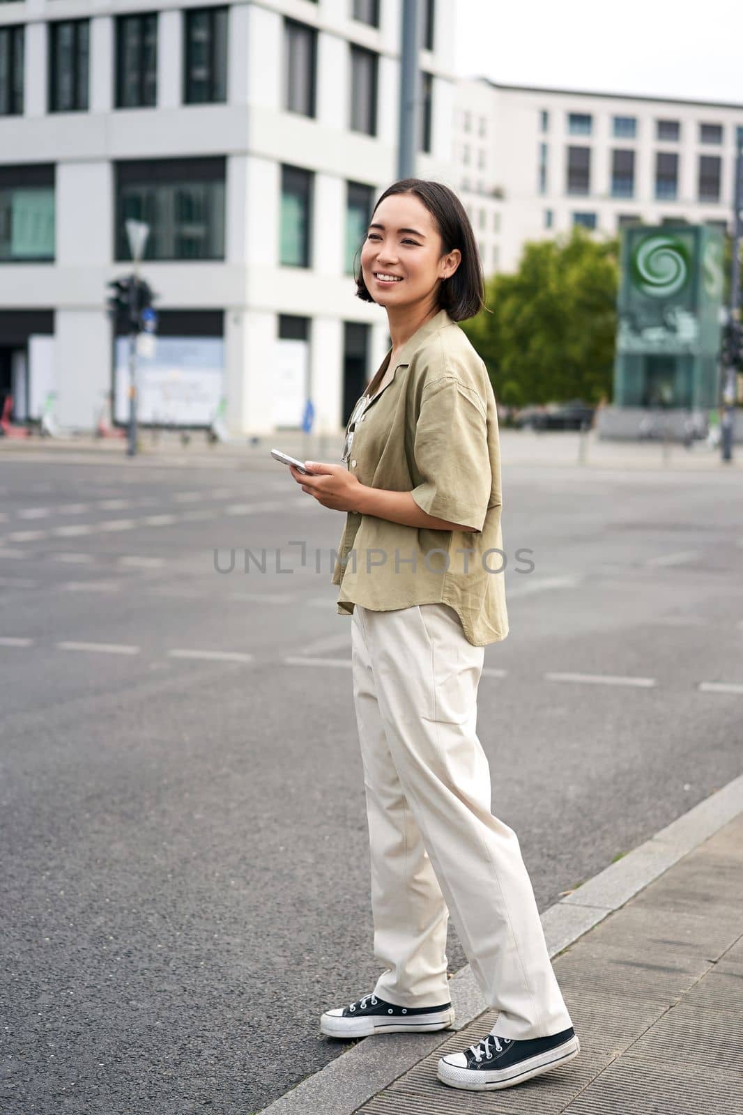 Car sharing and technology. Young asian woman waiting for taxi near road, holding smartphone, order car in application.