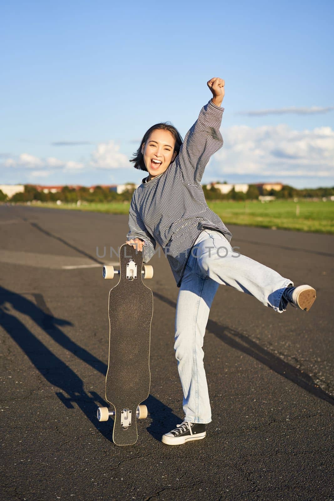 Vertical shot of asian girl feeling excited, skating on longboard, jumping and posing with skateboard, standing with cruiser on empty road, having fun outdoors by Benzoix