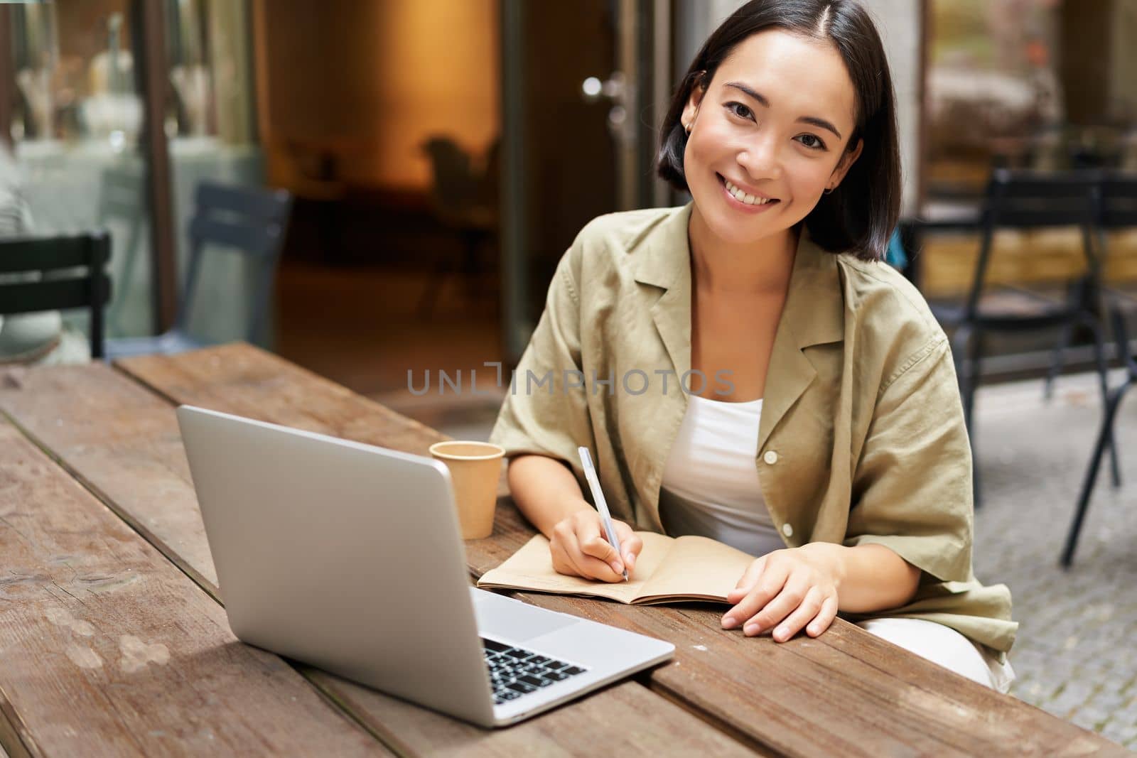 Portrait of young woman sitting in cafe with laptop, making notes, smiling at camera, working, having online meeting.