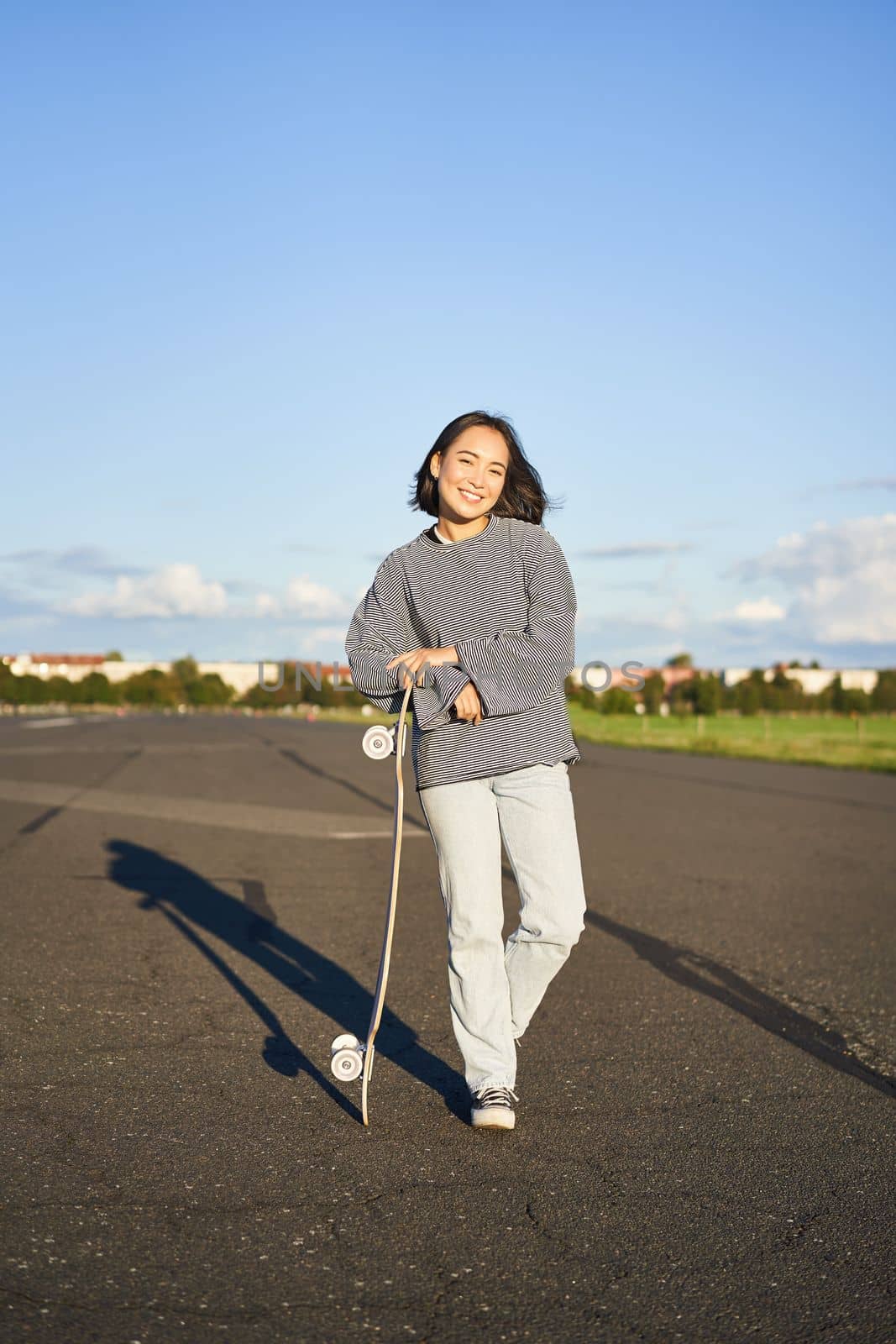 Vertical portrait of smiling asian woman standing on road with longboard, skateboarding on long cruiser, posing on empty road on sunny day.